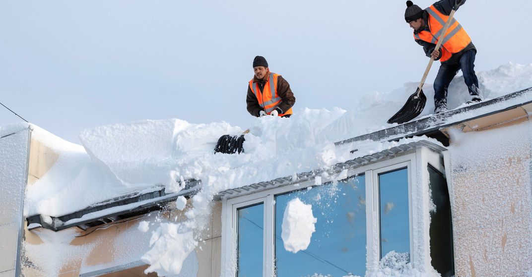 zwei Männer in orangen Warnwesten schaufeln mit Schneeschaufeln meterhohe Schneemassen von einem Dach