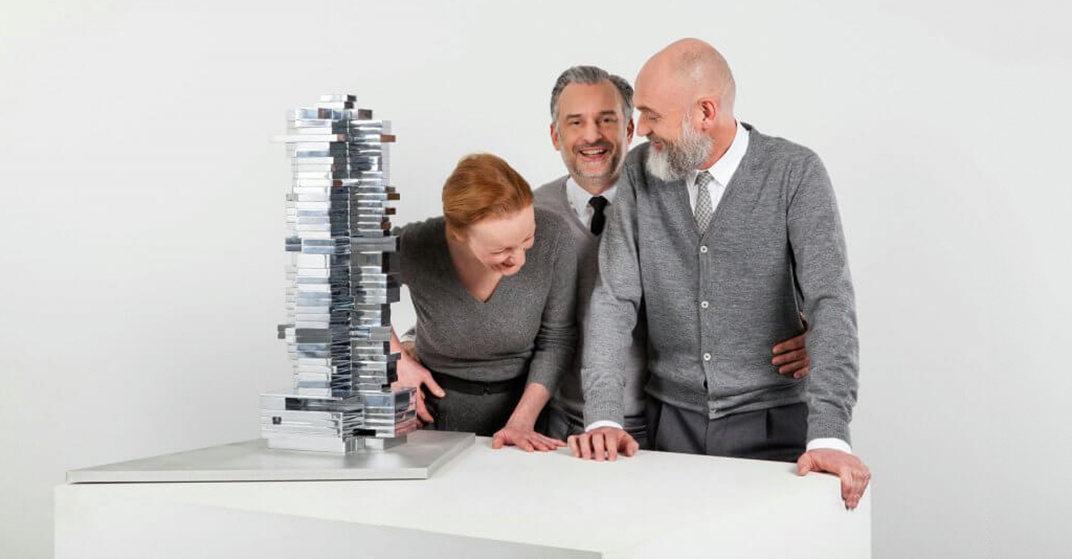 2 Männer und 1 Frau, alle in graue, elegante Pullover gekleidet, stehen lachend vor einem weißen Tisch mit einem silbernen Architekturmodell eines Hochhauses darauf. Führungsetage von BEHF Architects.