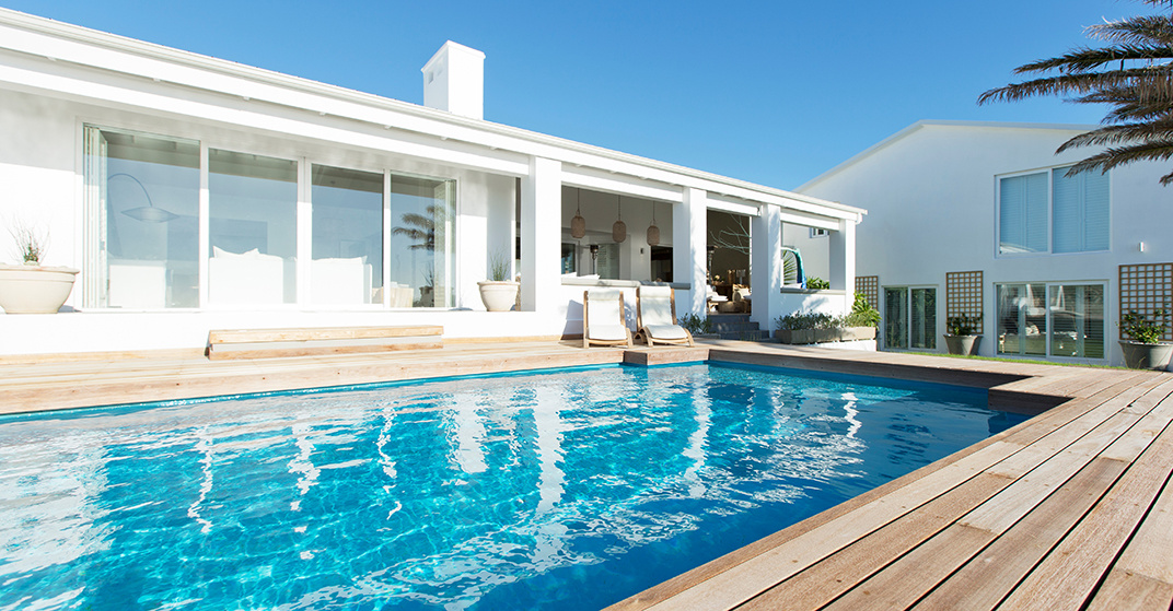 Weißes Haus mit Holzdeck und darin eingelassenen, sommerlichen Swimming Pool.