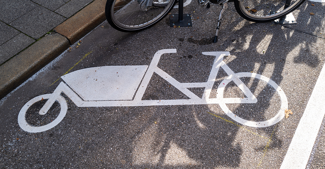 Parkplatzkennzeichnung Lastenrad auf städtischer Parkfläche. Sonne wirft Schatten, Fahrräder im Hintergrund zu Erkennen.