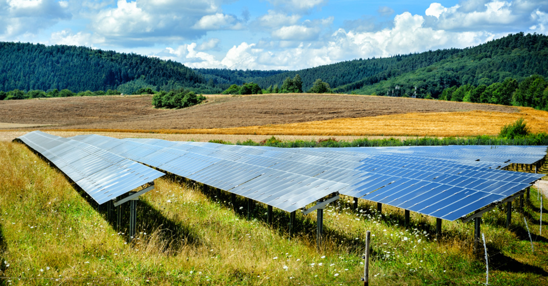 großer Solarpark auf grüner Wiese in hügeliger bewaldeter Landschaft unter blauem Himmel 