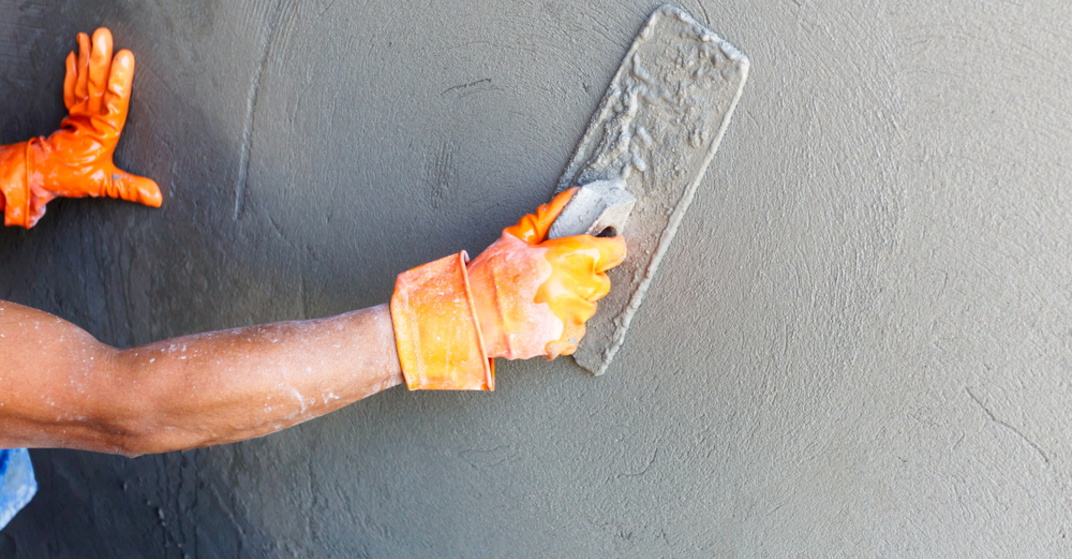 Mann mit orangen Arbeitshandschuhen verputzt eine Innenwand mit Maurerspachtel. Putz ist dunkelgrau und noch feucht. Ebene Oberfläche, wohl Grundputz.